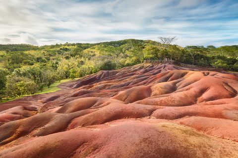 Colorful rocks and earth with shades of red, brown, and purple, surrounded by lush green trees under a partly cloudy sky.