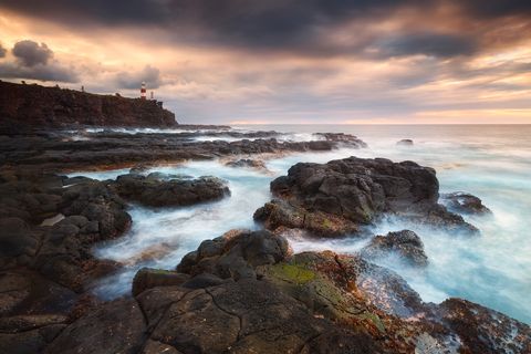 Rocky African coastline with dark volcanic rocks, gentle ocean waves, and a red-and-white Albion Lighthouse on a cliff under a dramatic sunset sky.