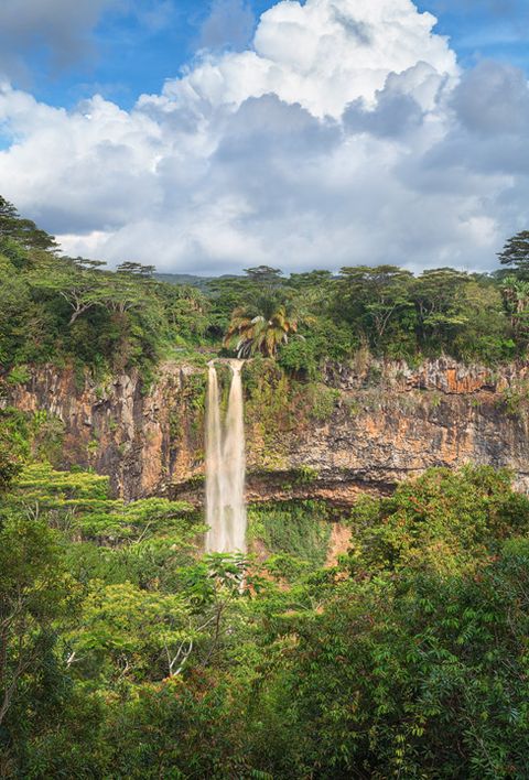 Tall waterfall cascading over a rocky cliff surrounded by lush green tropical forest under a partly cloudy sky.