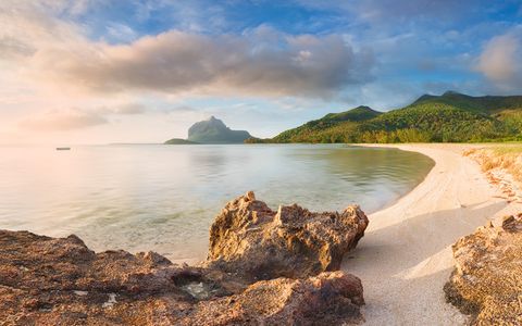 Tropical beach with golden sand, rocky foreground, calm sea, green mountains, and cloudy blue sky.