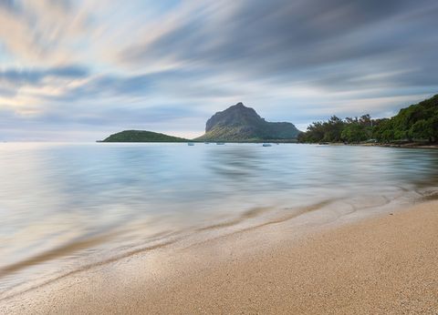 Sandy beach with calm water, distant green mountain, trees, and a dramatic cloudy sky.