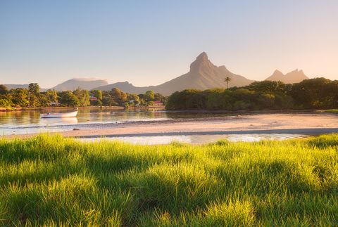 Peaceful riverside landscape with lush green grass, a small boat on calm water, and dramatic mountains in the background under warm sunlight.
