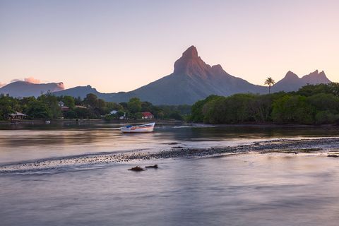Boat floating on calm river with lush greenery, houses, and a tall mountain peak in the background at sunset.