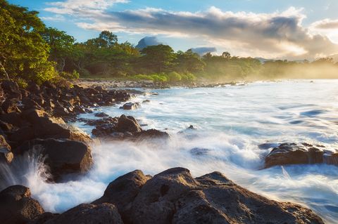 Waves crashing over dark volcanic rocks on a scenic tropical shoreline with lush green trees and cloudy sky at sunset.