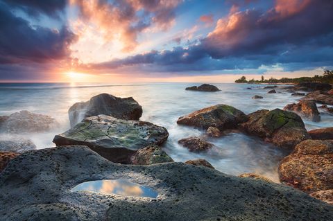 Rocky coastline at sunset with colorful clouds, calm ocean waves, and a small tide pool in the foreground.