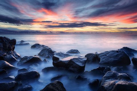 Large dark rocks emerging from calm ocean water at sunset with a colorful sky and dramatic clouds on Mauritius.