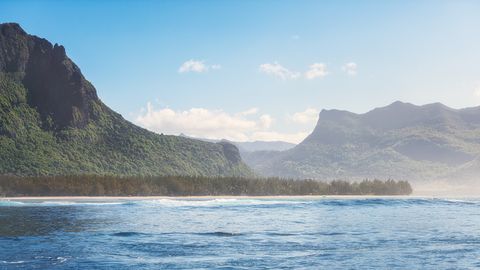Tropical coastline with blue ocean waves, dense green forest, and rugged mountain cliffs under a clear sky.