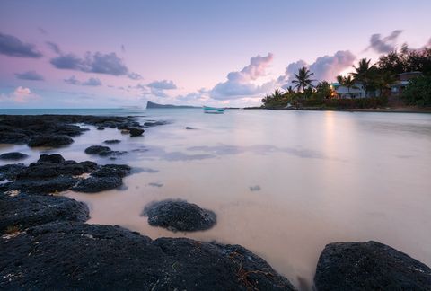 Tropical beach at sunset with black volcanic rocks in the foreground, calm water, palm trees, a small boat, and houses along the shoreline.