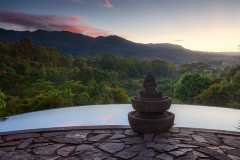 Stone statue on a pedestal overlooking an infinity pool with lush green forest and mountains in the background at sunrise