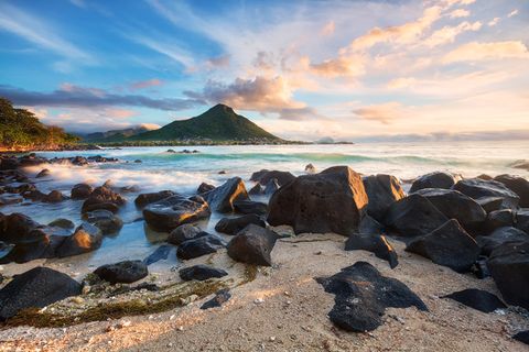 Rocky beach with large black stones, gentle waves, and a mountain under a colorful, cloudy sky at sunset.
