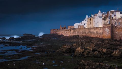 Essaouira city wall and white buildings overlooking rocky Atlantic coastline at dusk, dramatic sky in background.