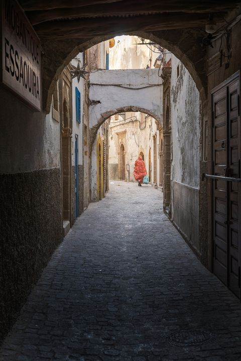 Narrow cobblestone alleyway in Essaouira, Morocco, with arched passageways, old weathered buildings, and a person in a red patterned cloak walking in the distance.