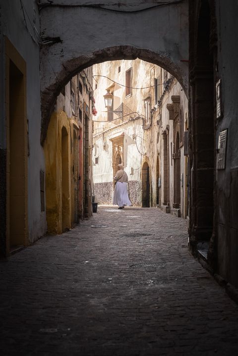 Schmale Kopfsteinpflastergasse in Essaouira, Marokko, mit alten, verwitterten Gebäuden und einer Person in traditioneller Kleidung, die unter einem Torbogen davongeht.