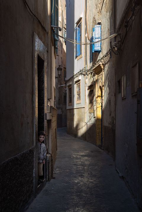 Narrow sunlit alley in Essaouira, Morocco, with aged walls, blue window shutters, and a young boy standing at a doorway in the shadows.