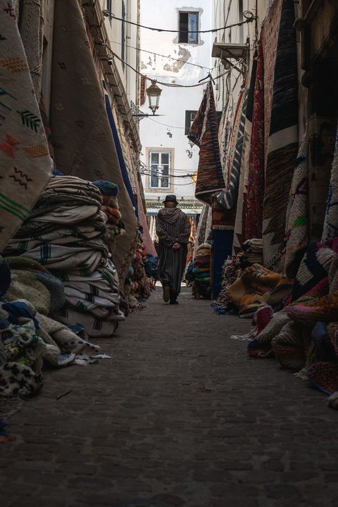 Narrow alleyway in Essaouira, Morocco, lined with colorful stacked carpets and rugs; a person in traditional clothing walks away in the center.
