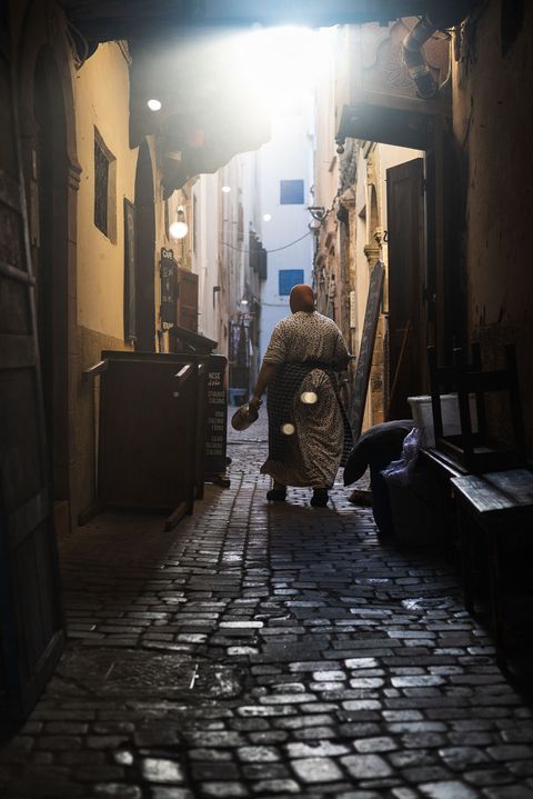 Person in traditional clothing walking down a narrow cobblestone alleyway in Essaouira, Morocco, with sunlight streaming in from above.