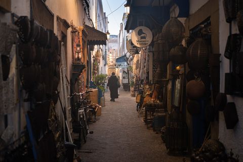 Narrow cobblestone alley in Essaouira, Morocco lined with vintage shops, traditional lanterns, carpets, and handicrafts, with a person walking in the distance.