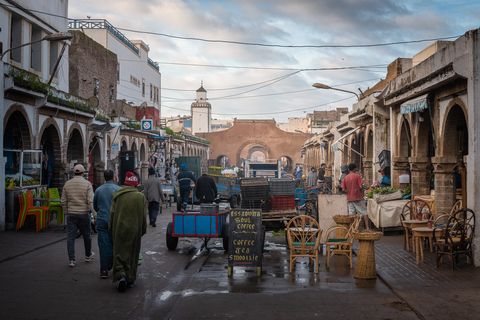 Street scene in Essaouira, Morocco with market stalls and a historic city gate and a mosque tower visible in the background under a cloudy sky.