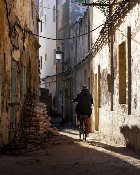 Person riding a bicycle through a narrow, sunlit alley with old, weathered buildings and stacked bags along the wall in Essaouira, Morocco.