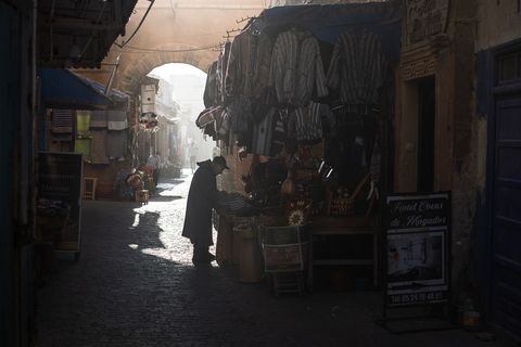 Narrow street market in Essaouira, Morocco with traditional clothing hanging, a person shopping in shadow, and sunlight streaming through an archway.