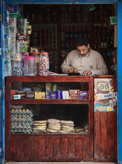 Small shop stall with a man standing behind a counter, displaying jars of sweets, chocolate bars, packs of eggs, and traditional Moroccan bread.