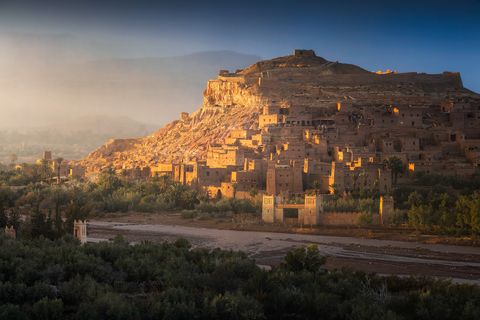 Ancient earthen buildings of Ait Benhaddou, a fortified village on a hill with desert landscape and greenery in Morocco at sunset.
