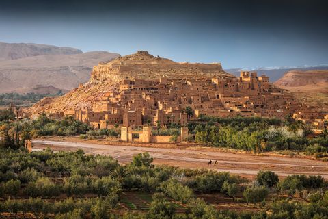 Ancient fortified village of Ait Benhaddou, Morocco, with traditional earthen clay buildings surrounded by green vegetation and arid mountains in the background.