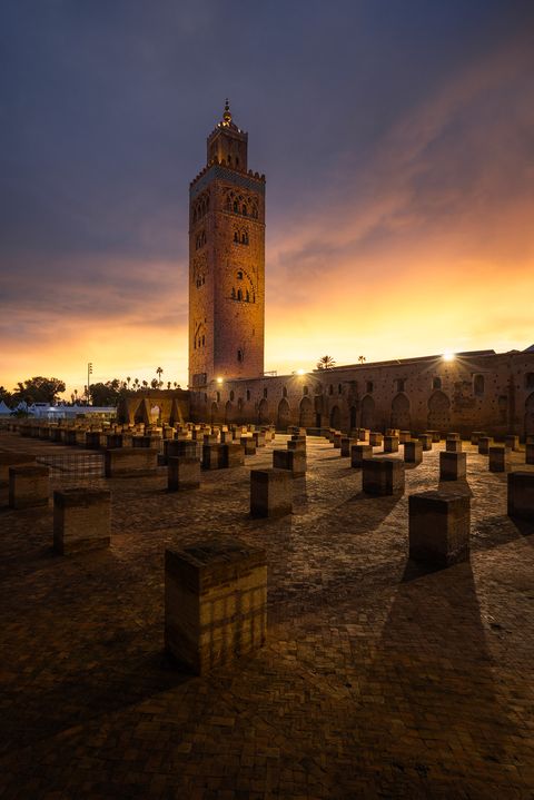 Koutoubia Mosque minaret in Marrakech, Morocco, illuminated at sunrise with a dramatic sky and stone pillars in the foreground.