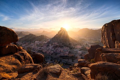 Sunrise over the traditional Moroccan village Agrd Odad, nestled among rocky mountains, with large reddish boulders in the foreground and Napoleon's Hat in the center.