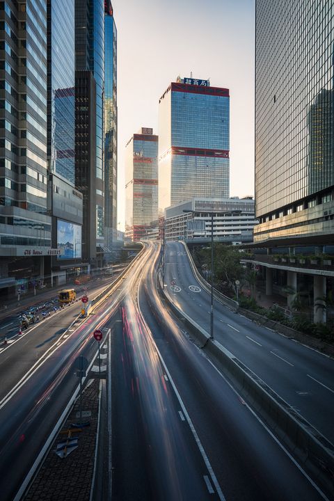 Cityscape of Hong Kong with tall glass skyscrapers and blurred light trails from moving vehicles on a multi-lane road at dusk.