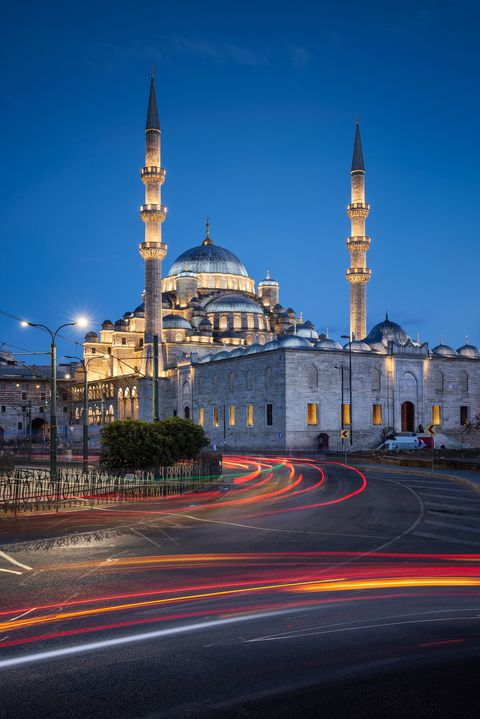 Illuminated mosque with two tall minarets at dusk in Istanbul, Turkey, with light trails from passing cars in the foreground.