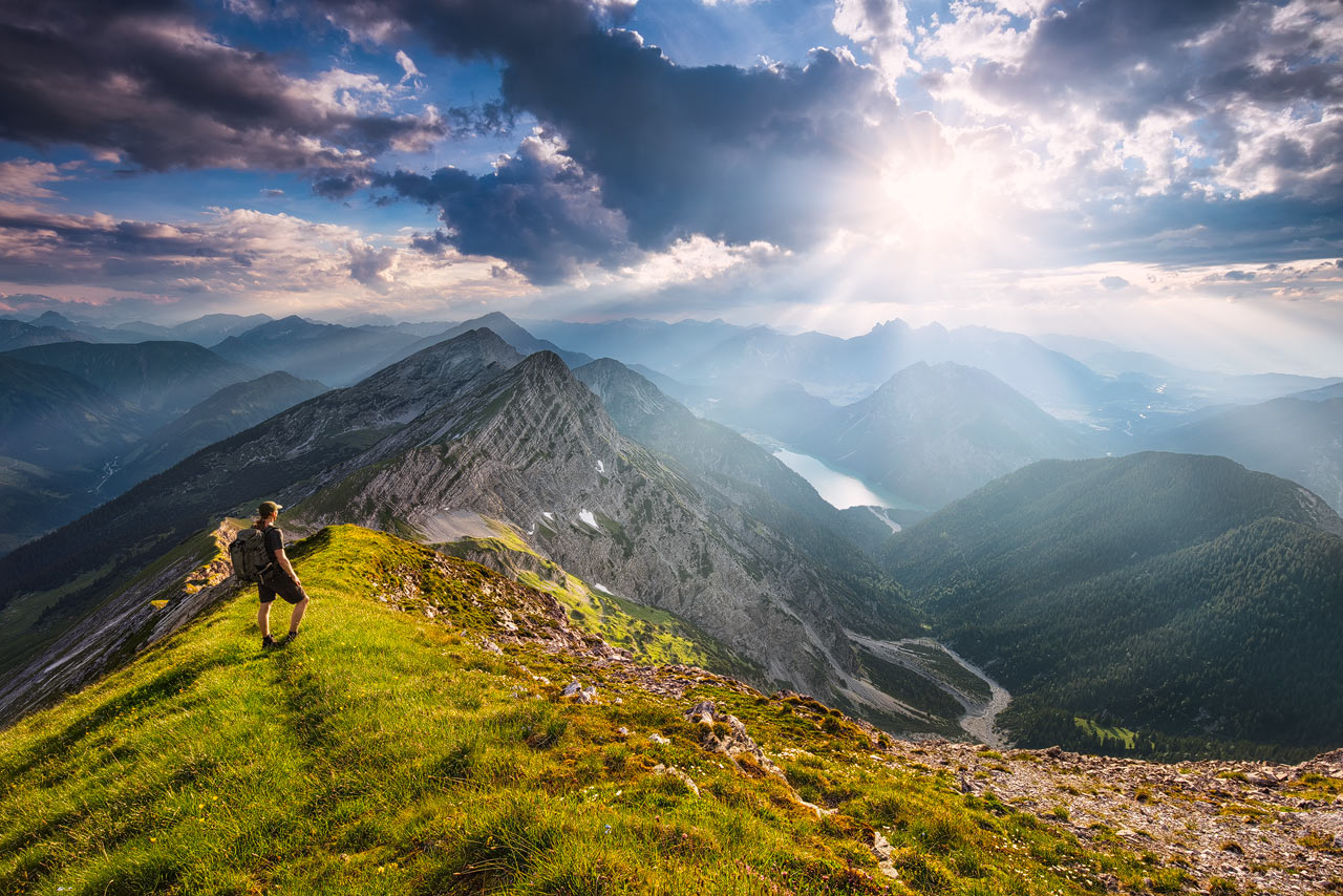 A hiker on a grassy ridge in the alps, looking toward teh Plansee.