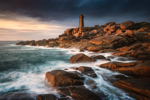 Rocky coastline with crashing waves and a lighthouse on top of the rocks at sunset in France.
