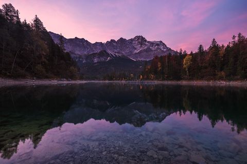 Alpine Eibsee lake with clear water reflecting the snow-capped Zugspitze mountain under a pink sunrise sky.