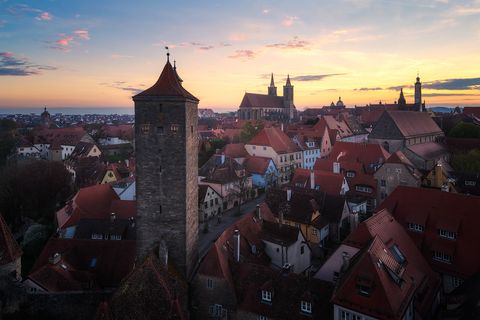 Medieval townscape at sunrise with red-tiled roofs, a prominent stone watchtower, historic buildings, and church spires in Rothenburg ob der Tauber, Germany.