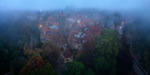 Aerial view of a medieval German town or Rothenburg shrouded in morning mist, featuring red-roofed buildings, stone towers, and surrounding trees.