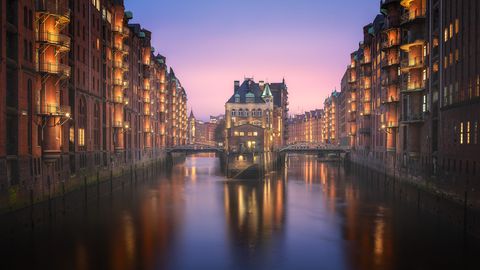 Historic red brick warehouses with illuminated balconies line both sides of a canal in Hamburg’s Speicherstadt district at dusk, with a central building on a small island and footbridges crossing the water.