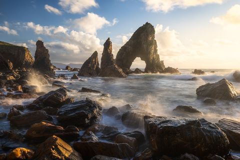Dramatic sea rocks and an arch formation at the coastline of Ireland at Crohy Head, with waves crashing against wet, dark boulders under a partly cloudy sky at sunset.