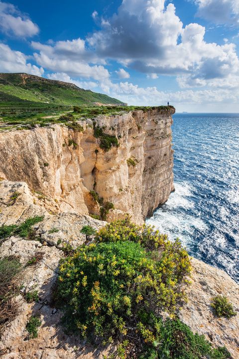 Steep Dingli cliffs with green vegetation overlooking a blue sea under a partly cloudy sky in Malta.