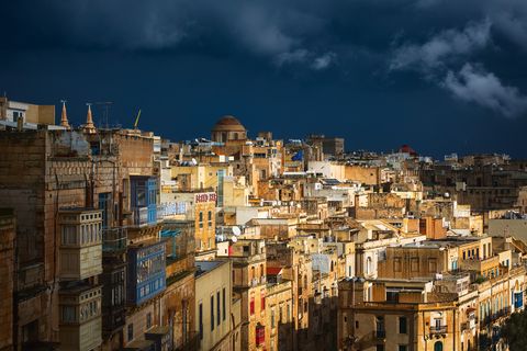 Historic cityscape of Valletta, Malta with limestone buildings, traditional colorful balconies, and dramatic stormy sky in the background.
