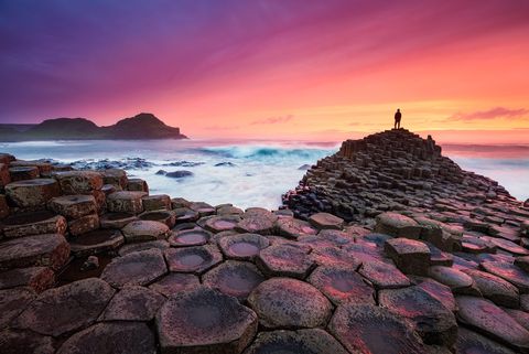 Giant’s Causeway at sunset with hexagonal basalt columns, a person standing on a rock formation, ocean waves, and a colorful sky in Northern Ireland.