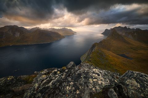 Dramatic view of a Norwegian fjord from the Barden Mountain with steep mountains, rugged cliffs, and dark storm clouds over deep blue water at sunset.