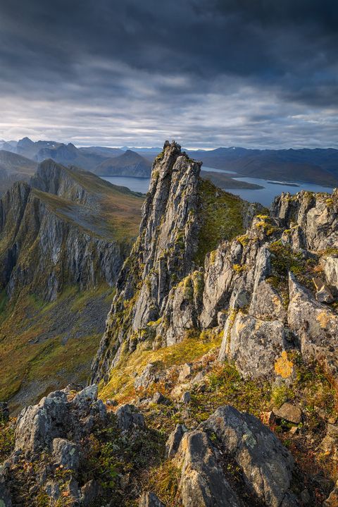 Sharp mountain ridge on the top of Husfjellet with rocky peaks and sparse vegetation, overlooking fjords and distant mountain ranges under a dramatic cloudy sky on Senja.