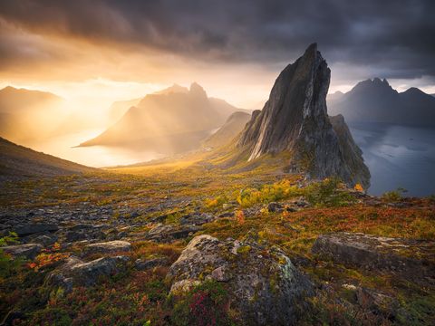 The Segla on Senja rising above a fjord at sunrise, with golden sunlight streaming through clouds and illuminating rocky terrain and autumn vegetation in Norway.
