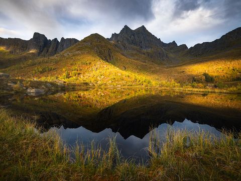 Dramatic mountain landscape on Senja with jagged peaks, grassy slopes, autumn colors, and clear lake reflecting the scenery under a partially cloudy sky.