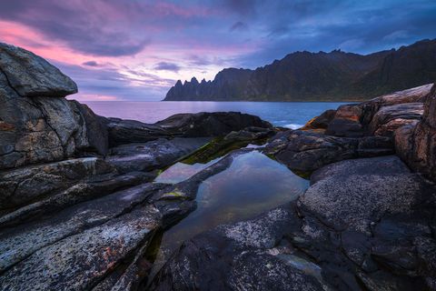 Rocky Norwegian coastline at Tungeneset at dusk with jagged mountains in the distance, tide pools reflecting the colorful sky, and calm sea.