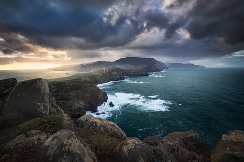 Dramatic coastal cliffs of Madeira with crashing waves, rocky shoreline, distant wind turbines, and a moody, cloudy sky at sunset.