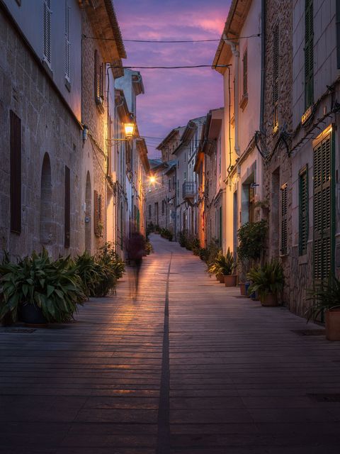 Narrow cobblestone street in Spain at dusk with old stone buildings and warm streetlights glowing under a purple sky.