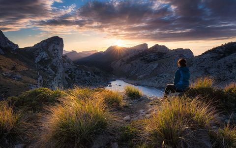 Person sitting on a grassy hilltop overlooking a mountain landscape of Cap Formentor at sunset.