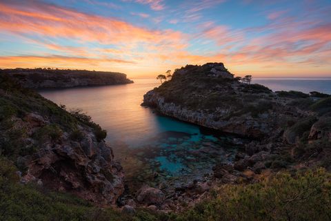 Rocky Cala des Moro with clear turquoise water, lush green vegetation, and a dramatic sunrise sky.